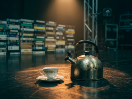A vintage tea kettle and porcelain teacup on an empty television studio stage floor in front of a wall of VHS tapes, evoking the Big Brother 28 tea party and Block Buster comp