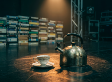 A vintage tea kettle and porcelain teacup on an empty television studio stage floor in front of a wall of VHS tapes, evoking the Big Brother 28 tea party and Block Buster comp