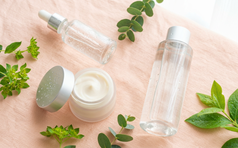 Dewy glass skin skincare products with a serum dropper, cream jar, and fresh green leaves