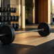 Rack of hexagonal rubber dumbbells and a loaded barbell on a wooden lifting platform in an empty gym lit by warm morning light