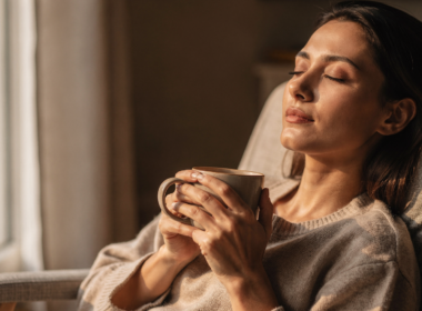 A person relaxing with eyes closed by a window holding a warm drink