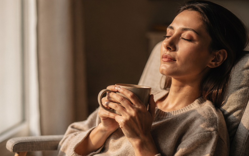 A person relaxing with eyes closed by a window holding a warm drink