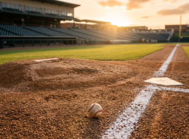 Empty baseball infield at dusk with freshly raked dirt and stadium lights coming on