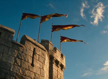 Low angle view of a medieval castle turret with crimson and gold pennants against a bright sky