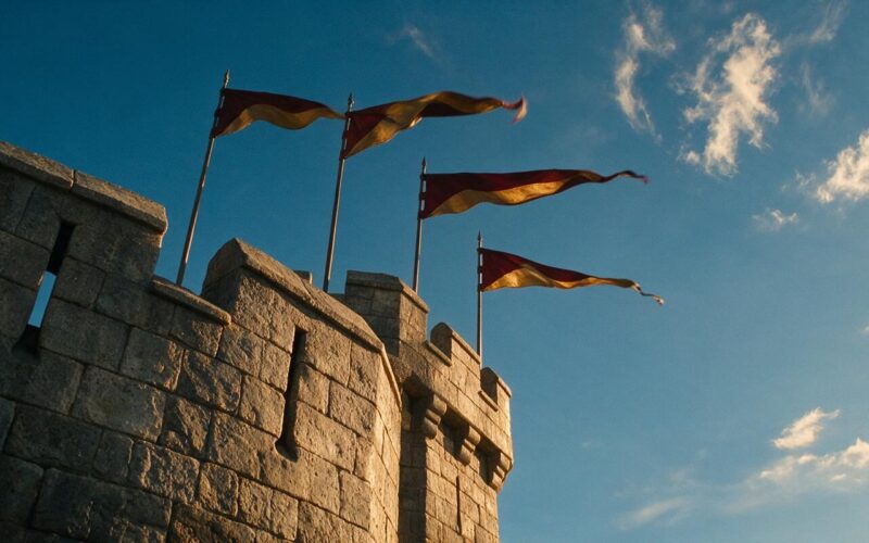 Low angle view of a medieval castle turret with crimson and gold pennants against a bright sky