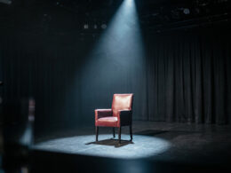 An empty game show stage at night with a single dramatic spotlight on one empty chair, evoking a tense Big Brother eviction