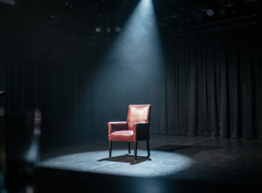 An empty game show stage at night with a single dramatic spotlight on one empty chair, evoking a tense Big Brother eviction
