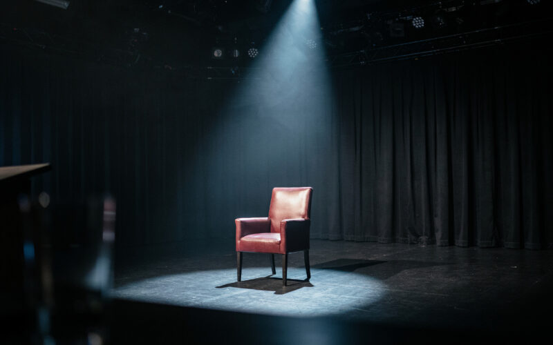 An empty game show stage at night with a single dramatic spotlight on one empty chair, evoking a tense Big Brother eviction