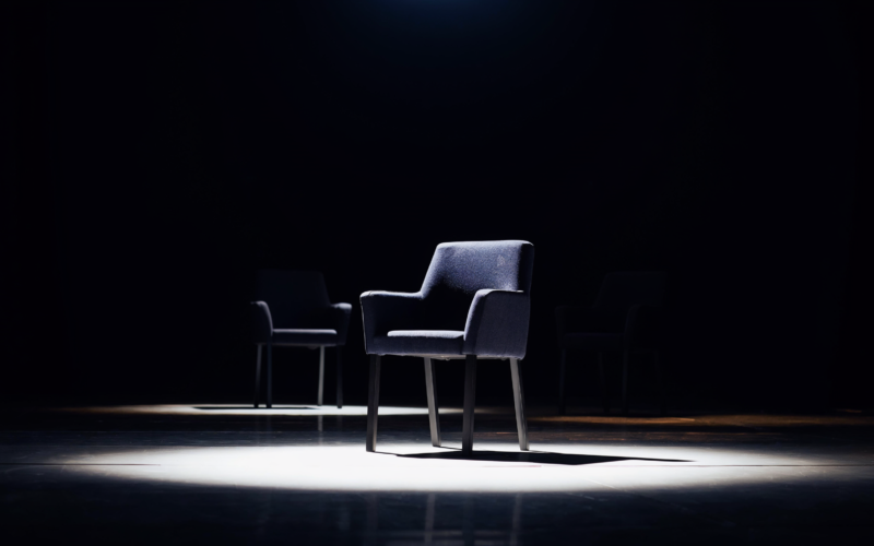 Big Brother 28 recap concept, single empty chair under a hard spotlight with two more in shadow