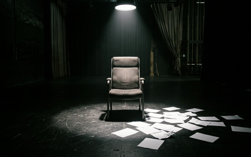Big Brother 28 eviction illustrated by an empty reality television interview chair under a hard spotlight with paper ballots scattered on a dark studio floor
