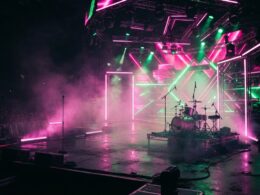 A vibrant neon lit concert stage with haze and pink and green lighting, evoking an energetic pop show