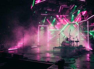 A vibrant neon lit concert stage with haze and pink and green lighting, evoking an energetic pop show