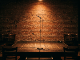 An empty comedy club stage with a single microphone stand under a warm spotlight and brick wall backdrop