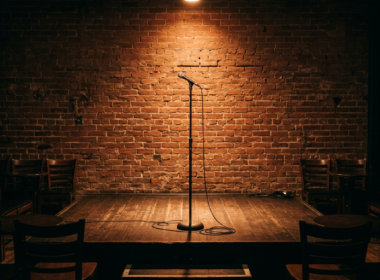 An empty comedy club stage with a single microphone stand under a warm spotlight and brick wall backdrop