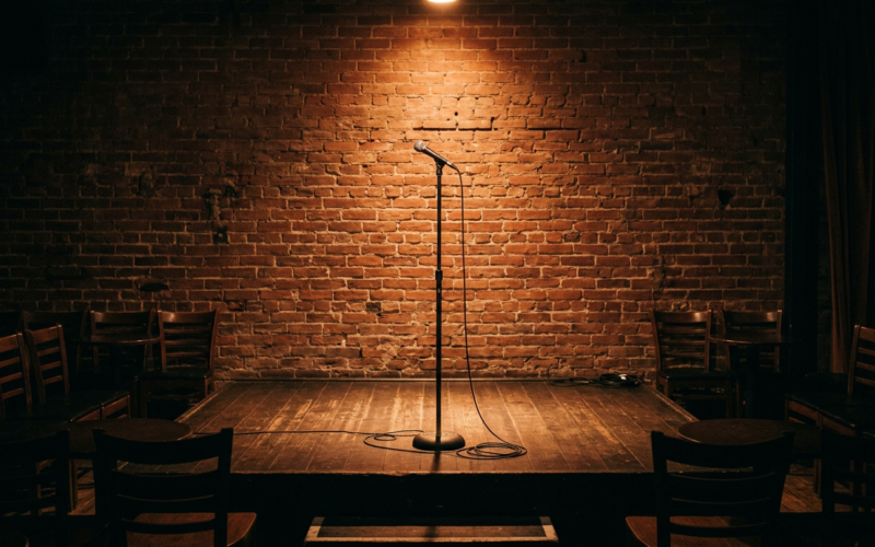 An empty comedy club stage with a single microphone stand under a warm spotlight and brick wall backdrop