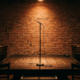 An empty comedy club stage with a single microphone stand under a warm spotlight and brick wall backdrop