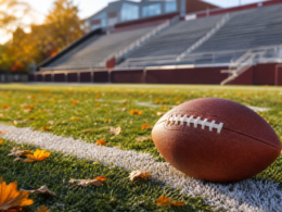 An American football resting on a chalk yard line of a sunlit college stadium field