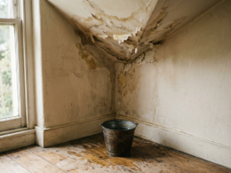 Home maintenance costs illustrated by a water stained ceiling corner with a metal bucket catching a drip on the floor below