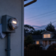 Residential electricity meter on a pale exterior wall at dusk with a folded utility statement tucked behind the conduit, illustrating 2026 electric bill increases