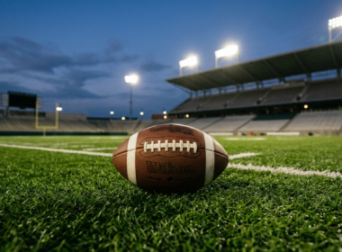 An American football resting on green stadium turf under bright lights at dusk