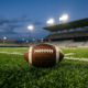 An American football resting on green stadium turf under bright lights at dusk