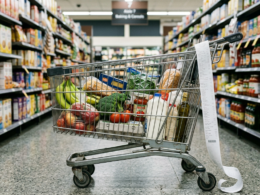 Half filled grocery cart in a supermarket aisle with a long receipt draped over the handle, illustrating 2026 food prices