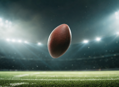 An American football spinning in mid air above green stadium turf under bright lights