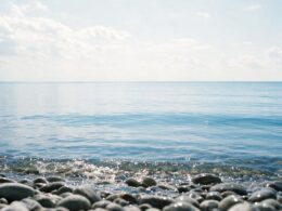 Low view across pebbles into a vast calm blue lake with an unbroken horizon and bright sky
