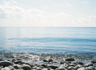 Low view across pebbles into a vast calm blue lake with an unbroken horizon and bright sky
