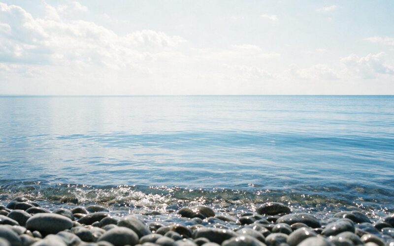 Low view across pebbles into a vast calm blue lake with an unbroken horizon and bright sky