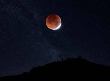 Deep red eclipsed moon with a bright white sliver above a dark desert ridge and star field