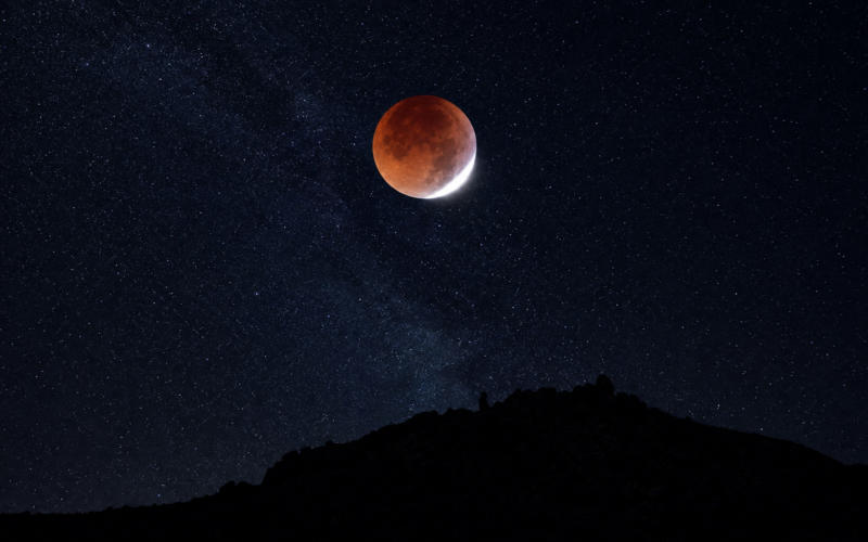 Deep red eclipsed moon with a bright white sliver above a dark desert ridge and star field