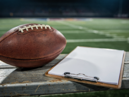An American football and a coaching clipboard on a sideline bench under stadium lights