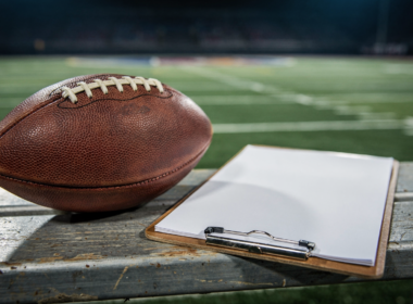 An American football and a coaching clipboard on a sideline bench under stadium lights