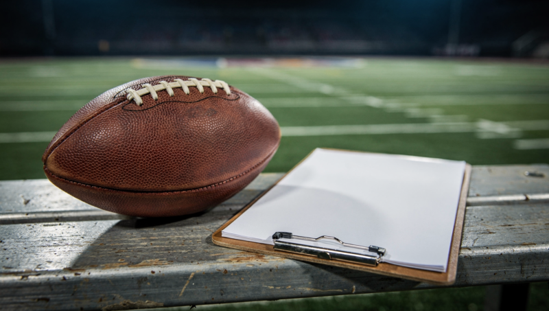 An American football and a coaching clipboard on a sideline bench under stadium lights