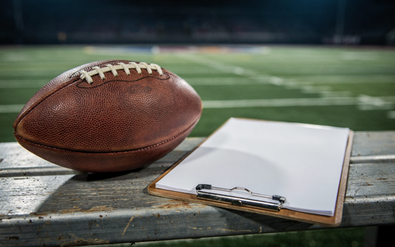 An American football and a coaching clipboard on a sideline bench under stadium lights