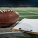 An American football and a coaching clipboard on a sideline bench under stadium lights