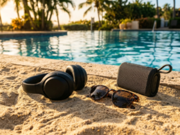 Headphones and sunglasses on bright sand beside a poolside speaker in golden summer light