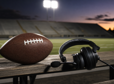 An American football and a coaching headset on a wooden sideline bench under stadium floodlights