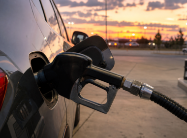 A gas station price sign glowing at dusk with an oil refinery silhouette in the distance